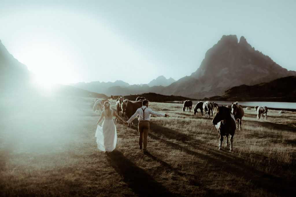 Photo d'un couple qui marche main dans les mains dans les montagnes des Pyrénées au lever de soleil. On y voit des chevaux et le pic du mid d'ossau. Une belle photo de fiançailles prise par la photographe de couple Gaétane Glize.
