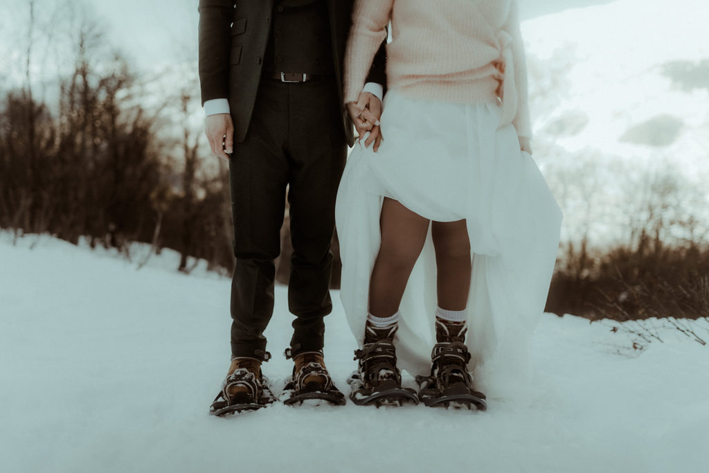 Photo détail d"un couple marié, on y voit leur tenues et des raquettes de neige à leurs pieds. Ils ont célébré leur mariage en faisant un elopement dans les montagnes des alpes. Un elopement d'hiver à la montagne et dans la neige. Photo de couple prise par Gaétane Glize, photographe de couple et de mariage.