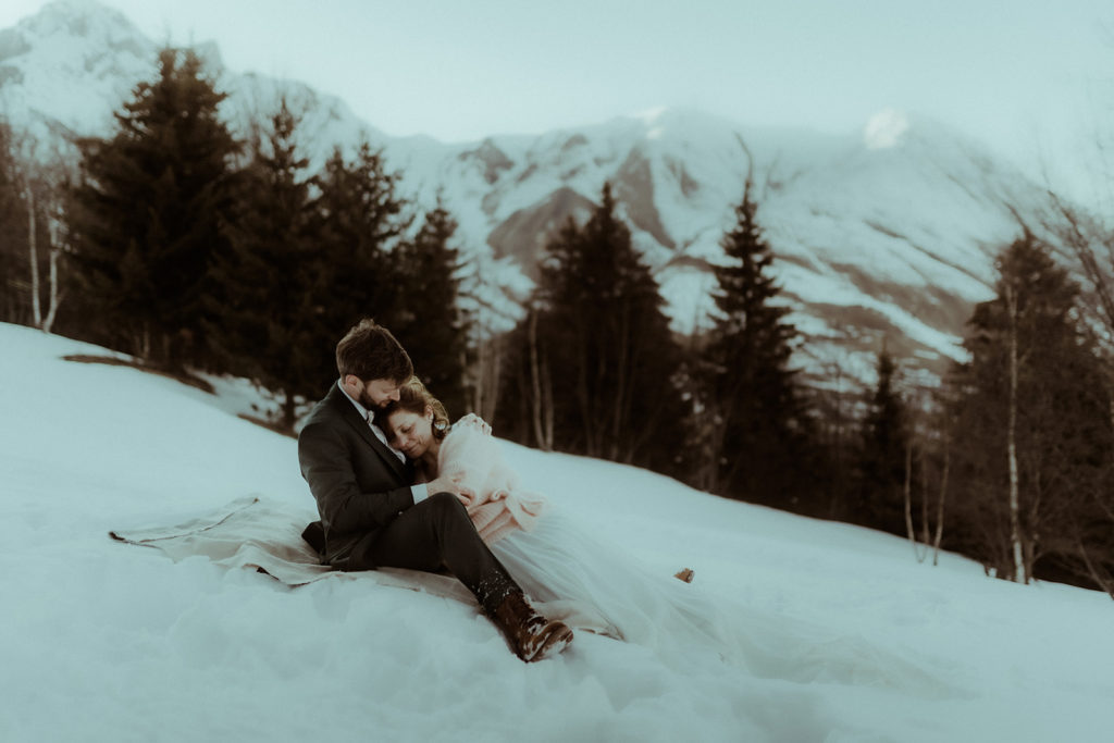 Photo d'un couple marié prise en montagne en hiver dans les Alpes. On y voit la neige, les sapins et les montagnes enneigées derrière. Photo prise par la photographe de mariage et de couple Gaétane Glize.