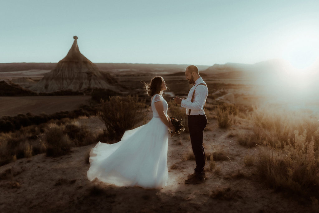 photo d'un couple marié dans leurs tenues de mariés avec la robe et le costume. Ils se lisent leurs voeux de mariage dans le désert des bardenas reales en espagne au coucher de soleil. Photo prise par la photographe de couple et de mariage Gaétane Glize.