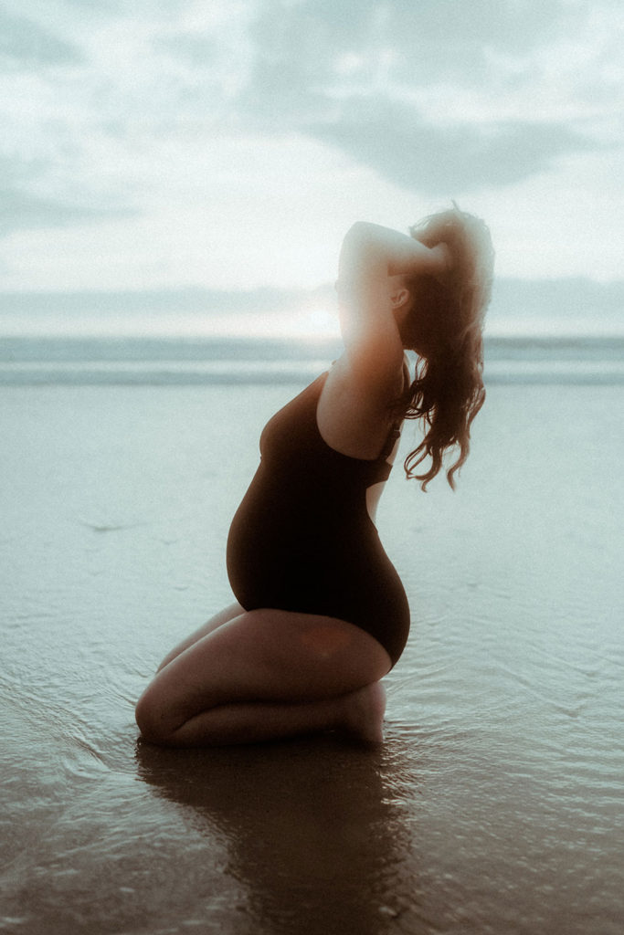 Femme enceinte assise dans l'eau de l'océan sur la plage de Seignosse dans les Landes au moment du coucher de soleil. Un joli moment capturé pendant sa séance photo grossesse.