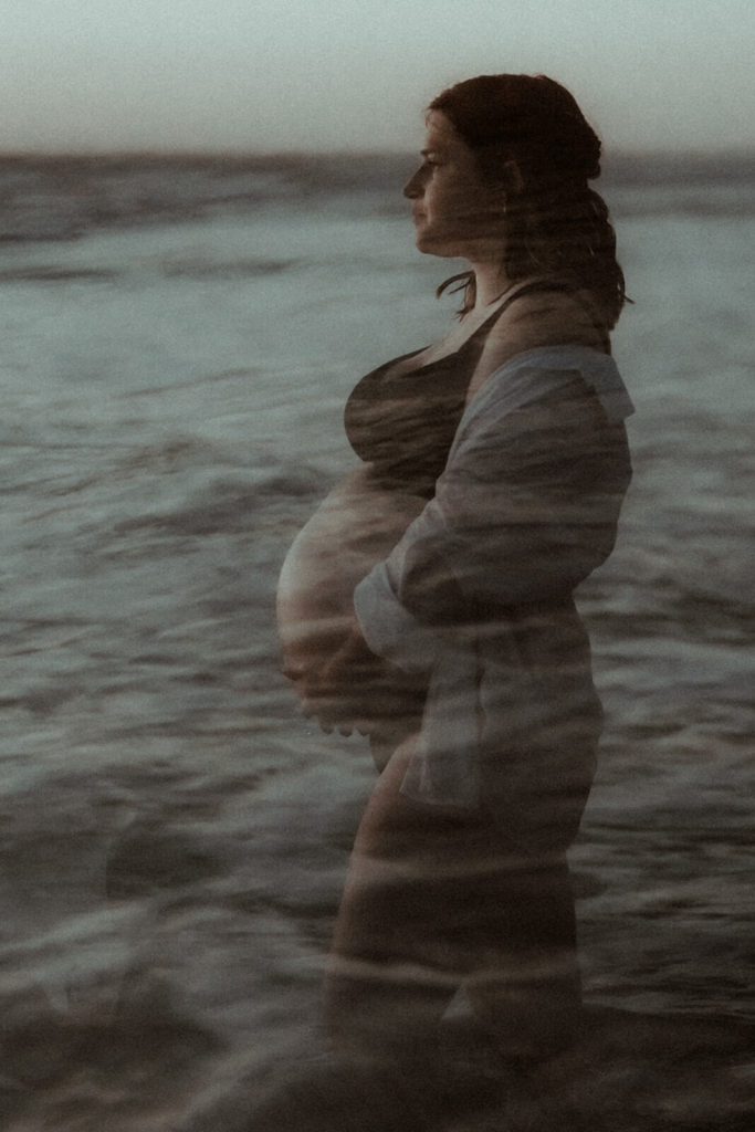 Séance photo grossesse sur la plage de Seignosse dans les landes au coucher de soleil. La femme enceinte est en sous-vêtements noirs avec une chemise qui dénude son épaule de façon très subtile et élégante. Elle est au bord de l'océan.