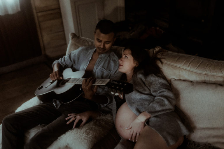 Un couple partage un moment de complicité dans un chalet en bois pendant leur séance photo de grossesse dans les landes. L'homme joue de la guitare pendant que la femme enceinte le regarde avec tendresse.