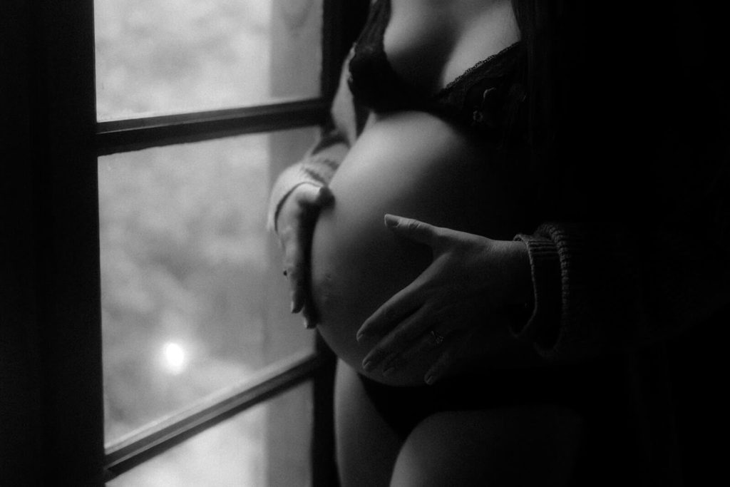 Photo gros plan en noir et blanc du ventre d'une femme enceinte en sous-vêtements noirs. Elle se tient debout près de la fenêtre à l'intérieur d'un chalet en bois dans les landes.
