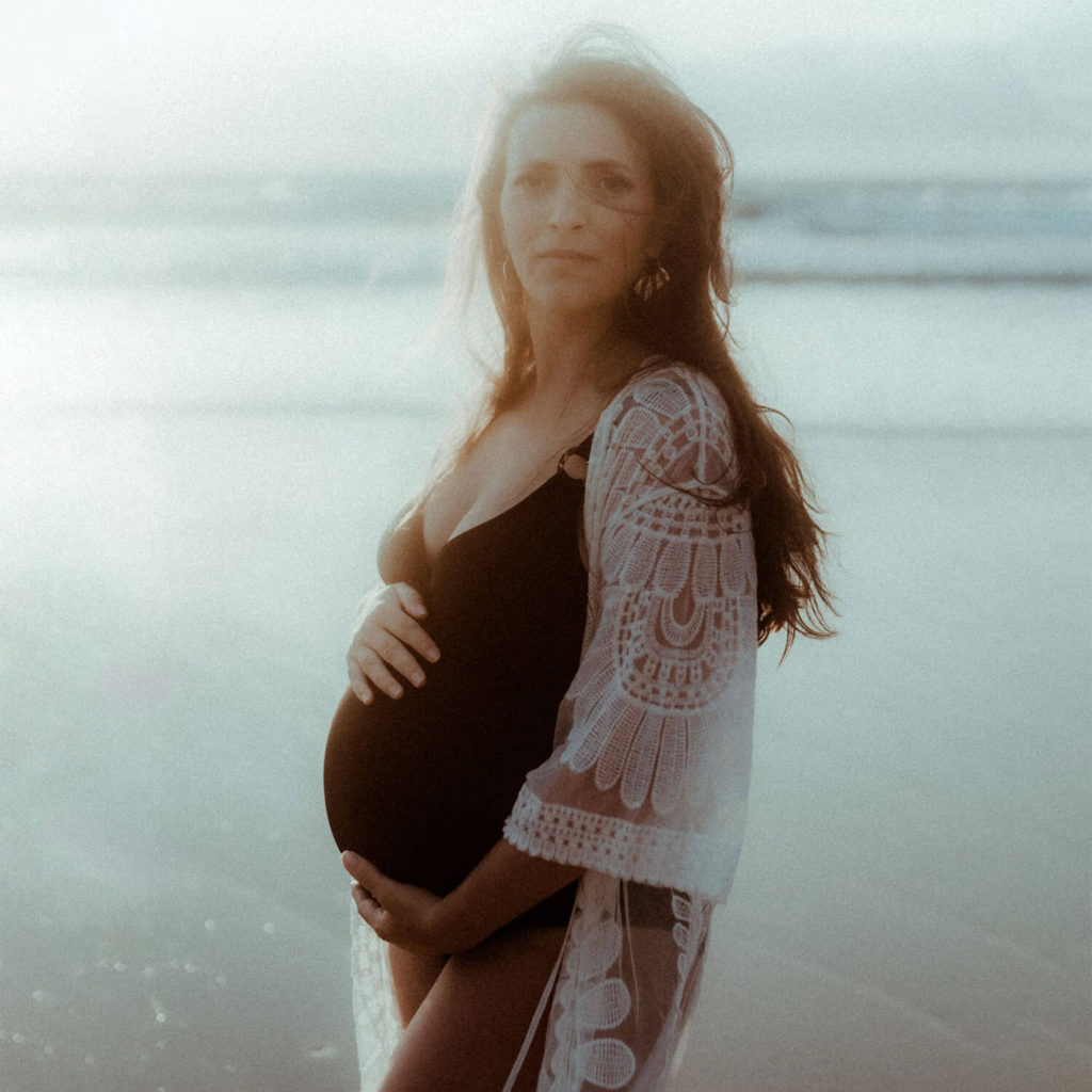 La femme enceinte pose ses mains sur son ventre et regarde droit devant au coucher de soleil. Une photo de grossesse prise par la photographe Gaétane Glize sur la plage à Seignosse dans les Landes.