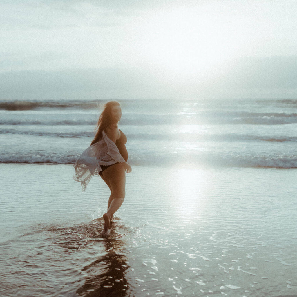 La femme enceinte marche dans l'eau de l'océan au coucher de soleil et se retourne pour regarder la photographe.